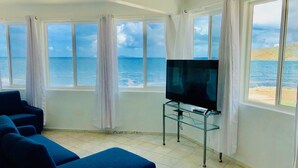 A room with a TV on a glass stand, a blue sofa, and a view of the sea through large windows. - The Waves; Oceanfront 2 Bedroom. Gorgeous Sunrises. Relax With The Wave Rythyms (Philipsburg)