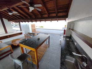 A covered outdoor area with a dining table, chairs, a small kitchen area, and a ceiling fan.