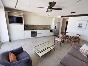 A modern kitchen with a built-in oven and refrigerator, a dining area with a glass table and chairs, and a ceiling fan.