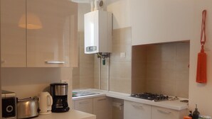 A compact kitchen with a gas stove, a coffee maker, and a toaster. There is a water heater mounted on the wall, and a bright pendant light hangs above the counter.