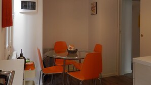 A dining area with a round table, four orange chairs, a hanging light fixture, a wall-mounted thermostat, and a door leading to another room.