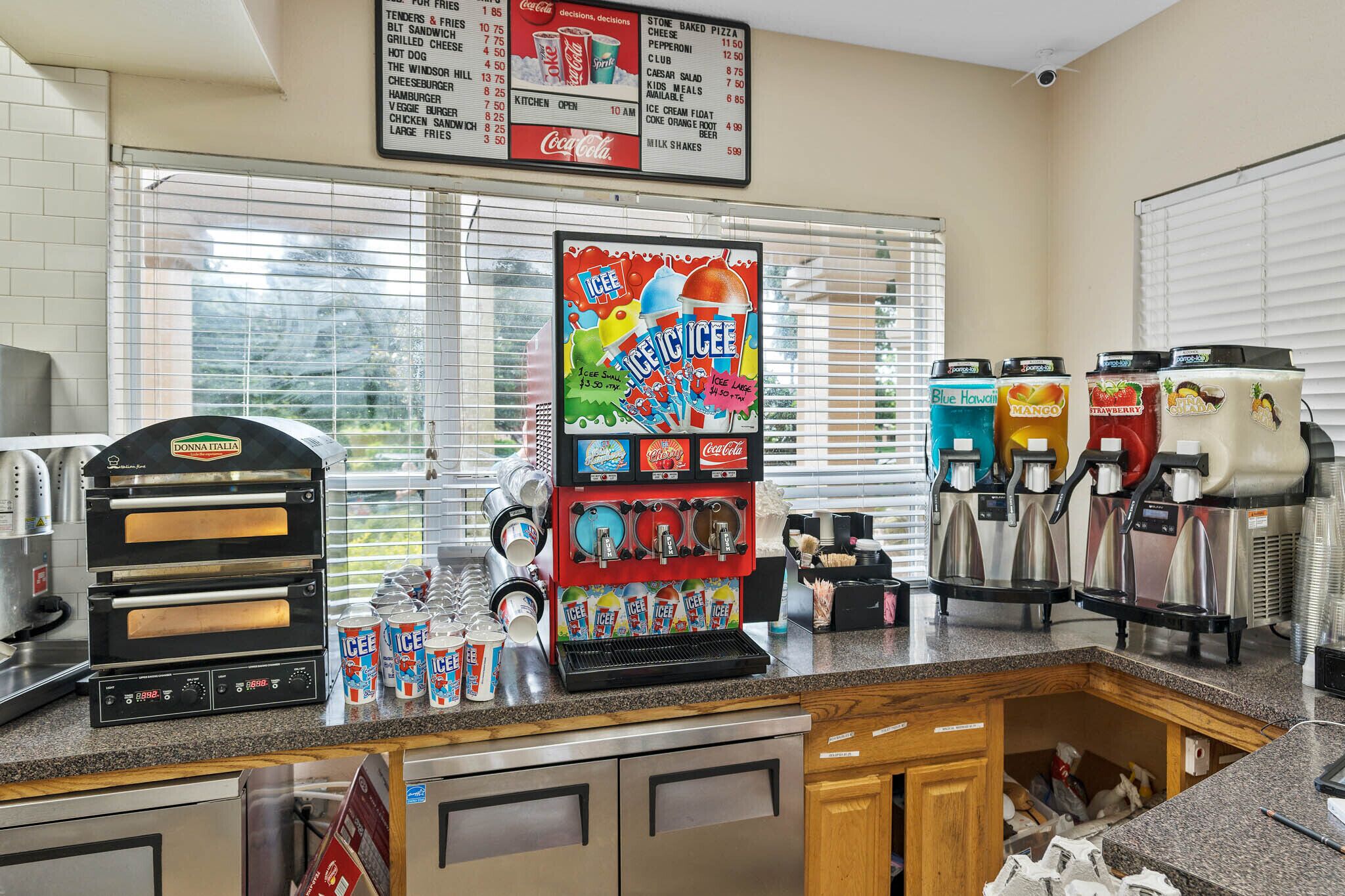 A kitchen with a menu board, a pizza oven, a beverage dispenser, and coffee machines.