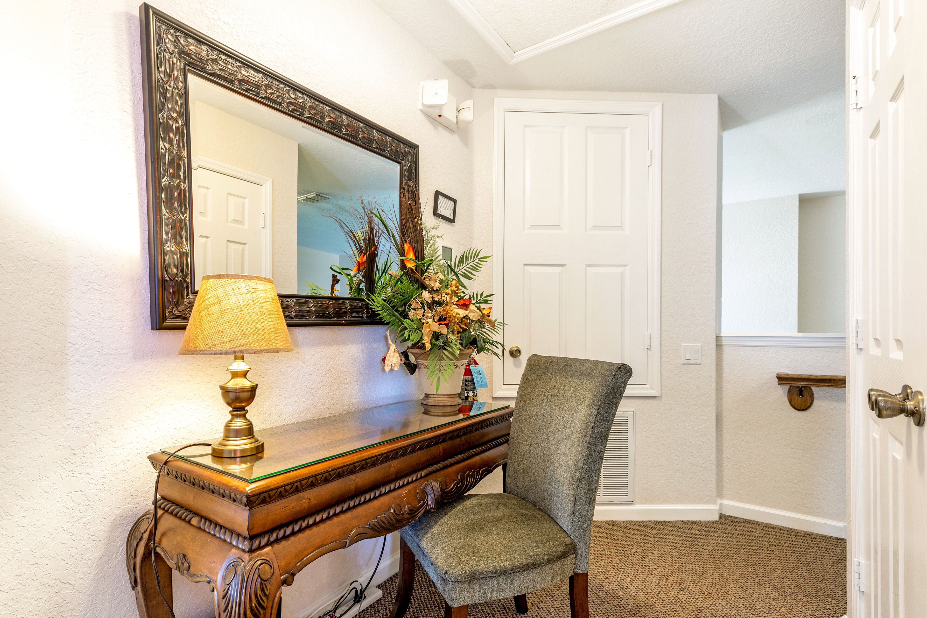 A corner with a wooden console table, a lamp, a chair, a mirror, and a vase with flowers.