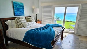 A bed with a blue blanket, a pillow with 'Sweet Dreams,' a lamp, and a view of the ocean through a door.