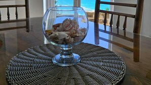 A dining table with a glass bowl of seashells, a woven placemat, and a view of the ocean through a door.