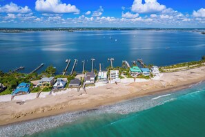 An aerial view of a coastal area with a beach, several piers, and buildings with a clear view of the ocean and distant islands.