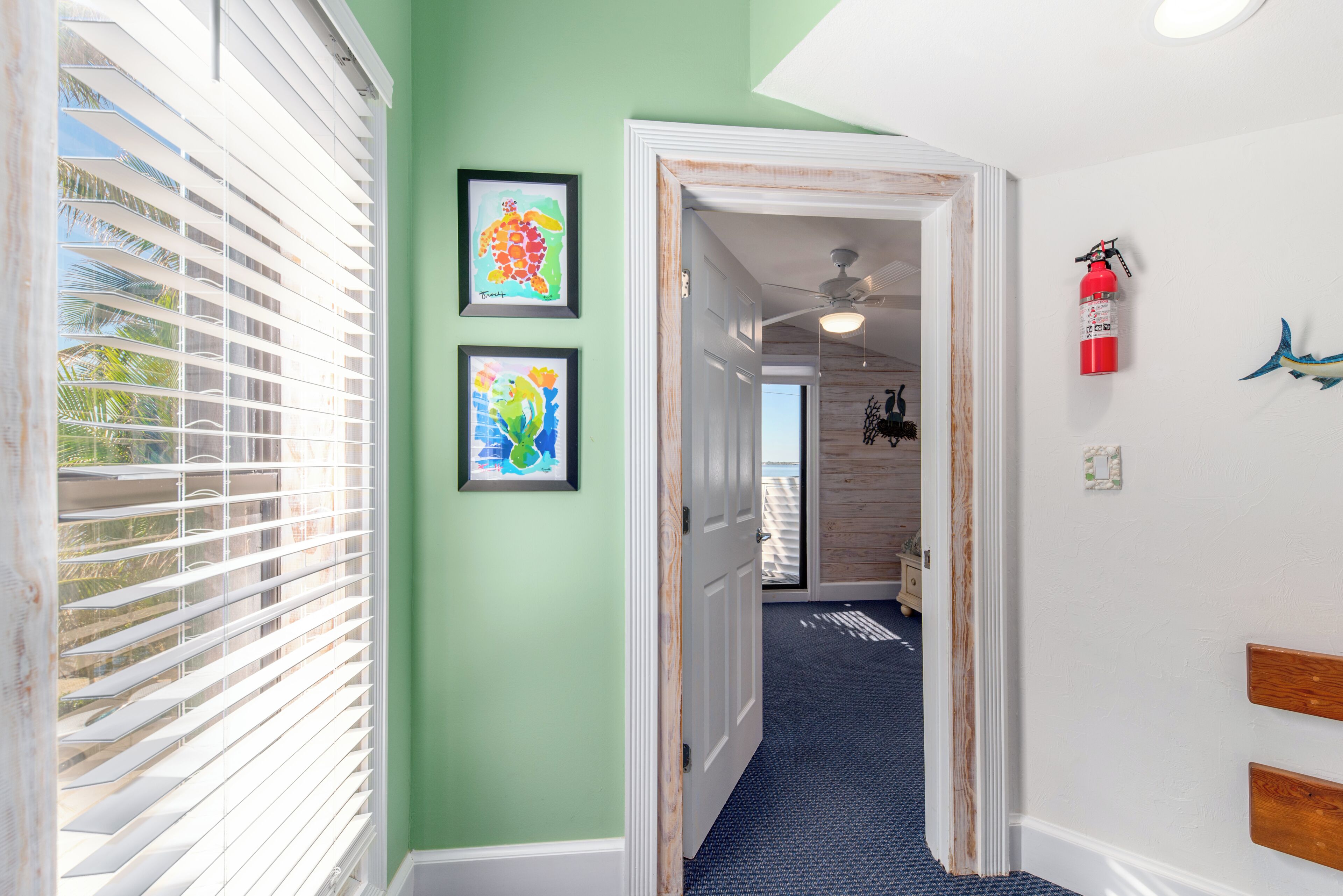 Un couloir aux murs verts, orné de deux œuvres d’art encadrées, avec un ventilateur de plafond et un extincteur.