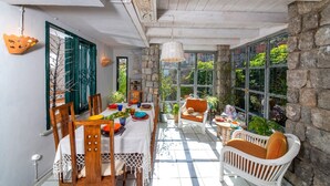 Un coin repas baigné de lumière, avec une table dressée pour deux, entouré de murs en pierre et de grandes fenêtres.