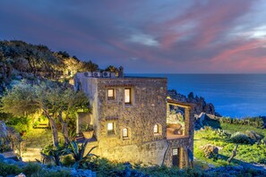 Un edificio de piedra con varias ventanas y balcones, rodeado de abundante vegetación y vistas a un cuerpo de agua.