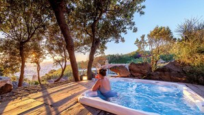 Une terrasse en bois avec un jacuzzi donnant sur une étendue d’eau, entourée d’arbres et de rochers.