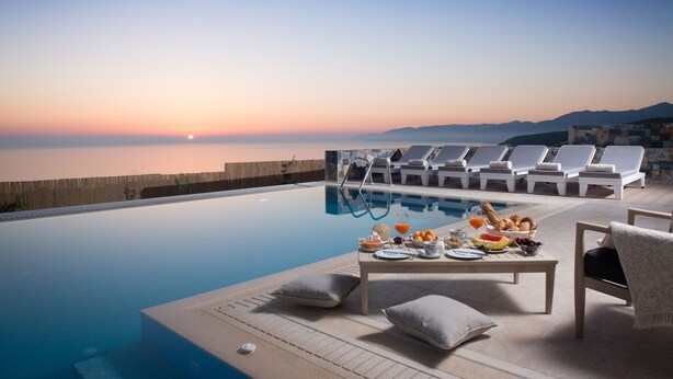A poolside area with lounge chairs and a table set for breakfast, overlooking a body of water and mountains.