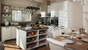 A modern kitchen with white cabinetry, a central island with baskets of fruits and vegetables, and a dining area with a white table set for two.