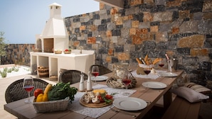 An outdoor dining area with a stone wall, a white barbecue, wicker chairs, and a table set with plates, glasses, and food.