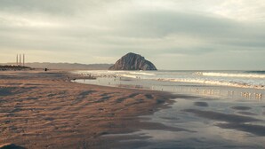 A beach with a large rock formation in the distance, waves approaching the shore, and a cloudy sky.