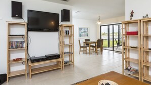 Un salon moderne doté d’un téléviseur à écran plat, d’étagères en bois, d’un coin repas avec une table et des chaises, et offrant une vue sur un lac grâce à de grandes fenêtres.