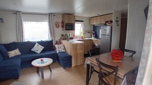 A compact living space with a sofa, a small round table, a dining table with chairs, a kitchenette, and a window with curtains. - Spacious, comfortable mobile home for 6/8 people (40m2) (Les Mathes)
