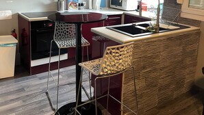 Une cuisine compacte au design moderne, dotée d’une hotte noire, d’armoires rouges, d’un four à micro-ondes et d’un petit îlot central avec un évier et des tabourets.