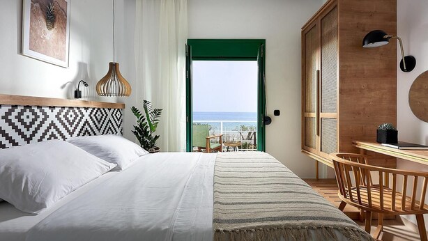 A bedroom with a bed, a desk, a chair, a plant, and a view of the sea through a green door. - Beach Villa in Crete - Villa Kirvas (Ierapetra)