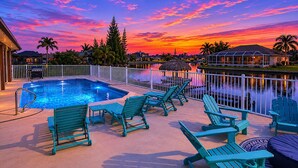 A swimming pool with lounge chairs, a view of a sunset over water, and a fenced area. - Villa Coconut Cove - Beautiful waterfront home w. heated pool (Cape Coral)