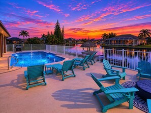 Une piscine avec des chaises longues, une vue sur un coucher de soleil sur l’eau et un espace délimité par une clôture.