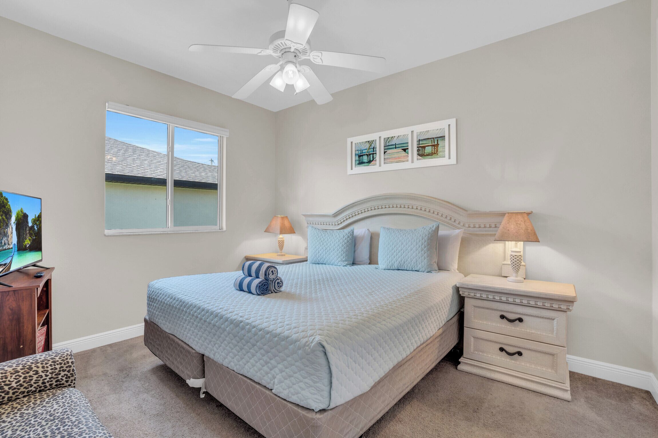A bedroom with a bed, nightstands, a ceiling fan, a window with a view, and artwork on the wall.
