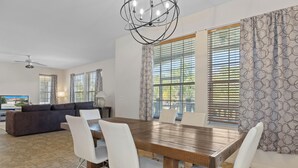 A modern dining area with a wooden table and white chairs, large windows with blinds, a ceiling fan, and a dark-colored sofa in the background. - Waterfront! Boat dock available for rent, pet friendly, families welcome!  (Homosassa)