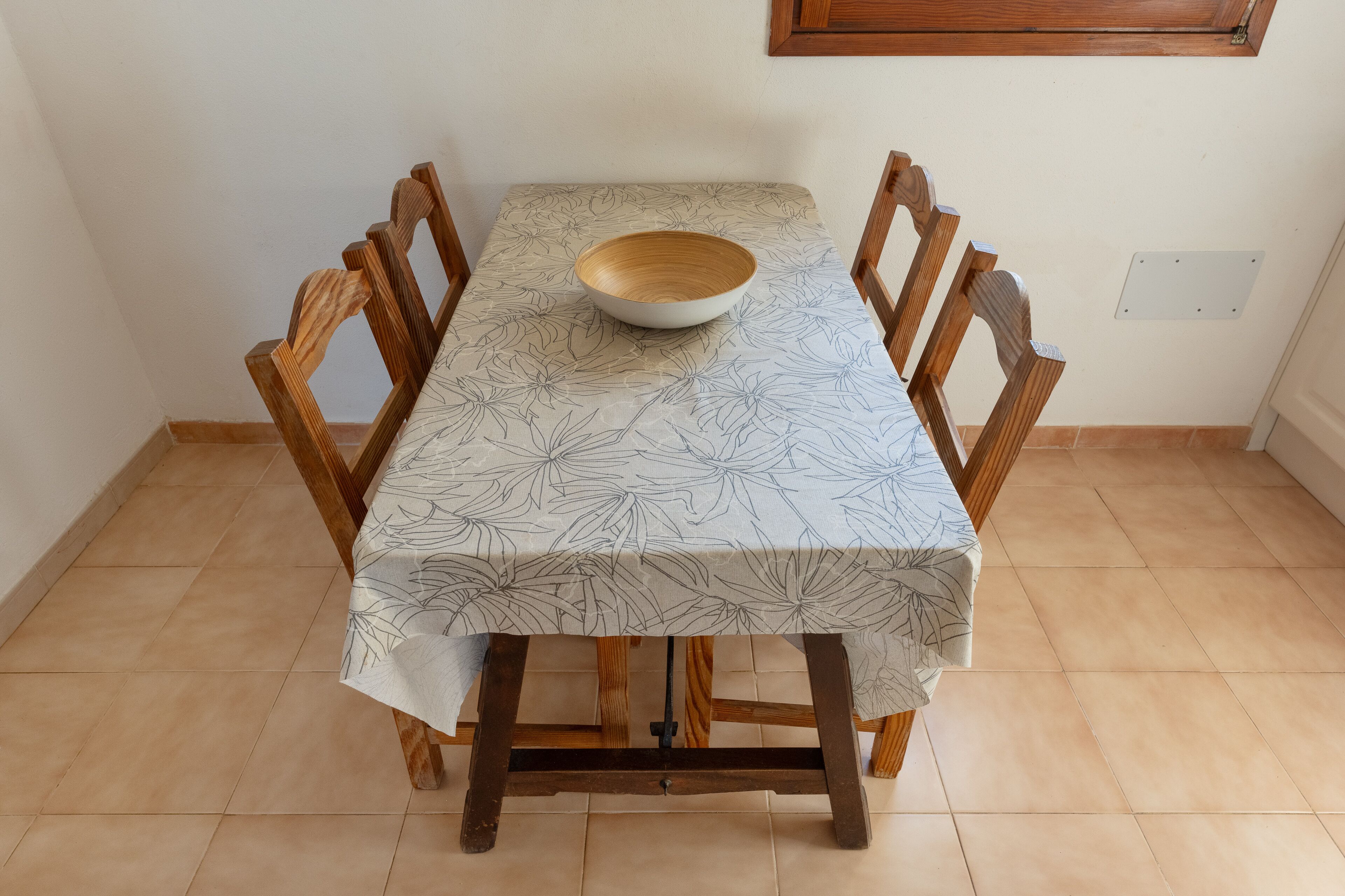 A dining table with a floral tablecloth, wooden chairs, and a wooden bowl on the table.
