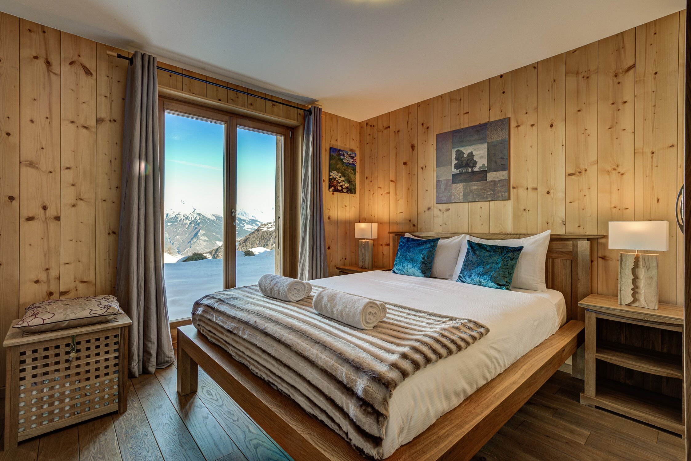 Une chambre aux murs lambrissés, avec un grand lit, deux lampes de chevet, une table de chevet et une vue sur les montagnes par une porte coulissante.