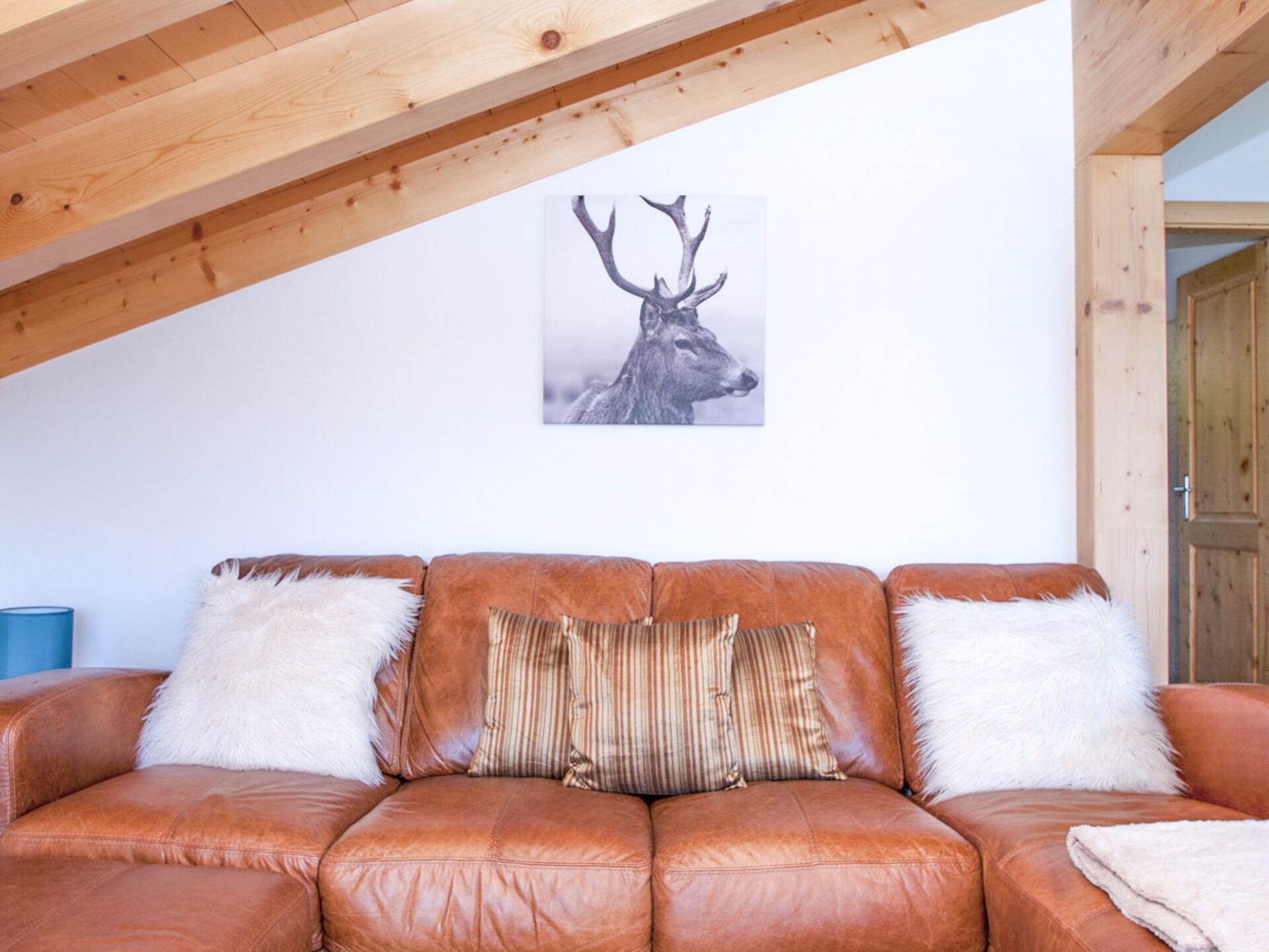 Un canapé en cuir marron agrémenté de coussins blancs et à rayures, une œuvre d’art représentant une tête de cerf encadrée au mur et un plafond en bois.