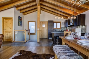 Une cuisine spacieuse avec des poutres en bois, un coin repas avec une table en bois dressée pour quatre personnes, et un grand chien allongé sur un tapis en fourrure.