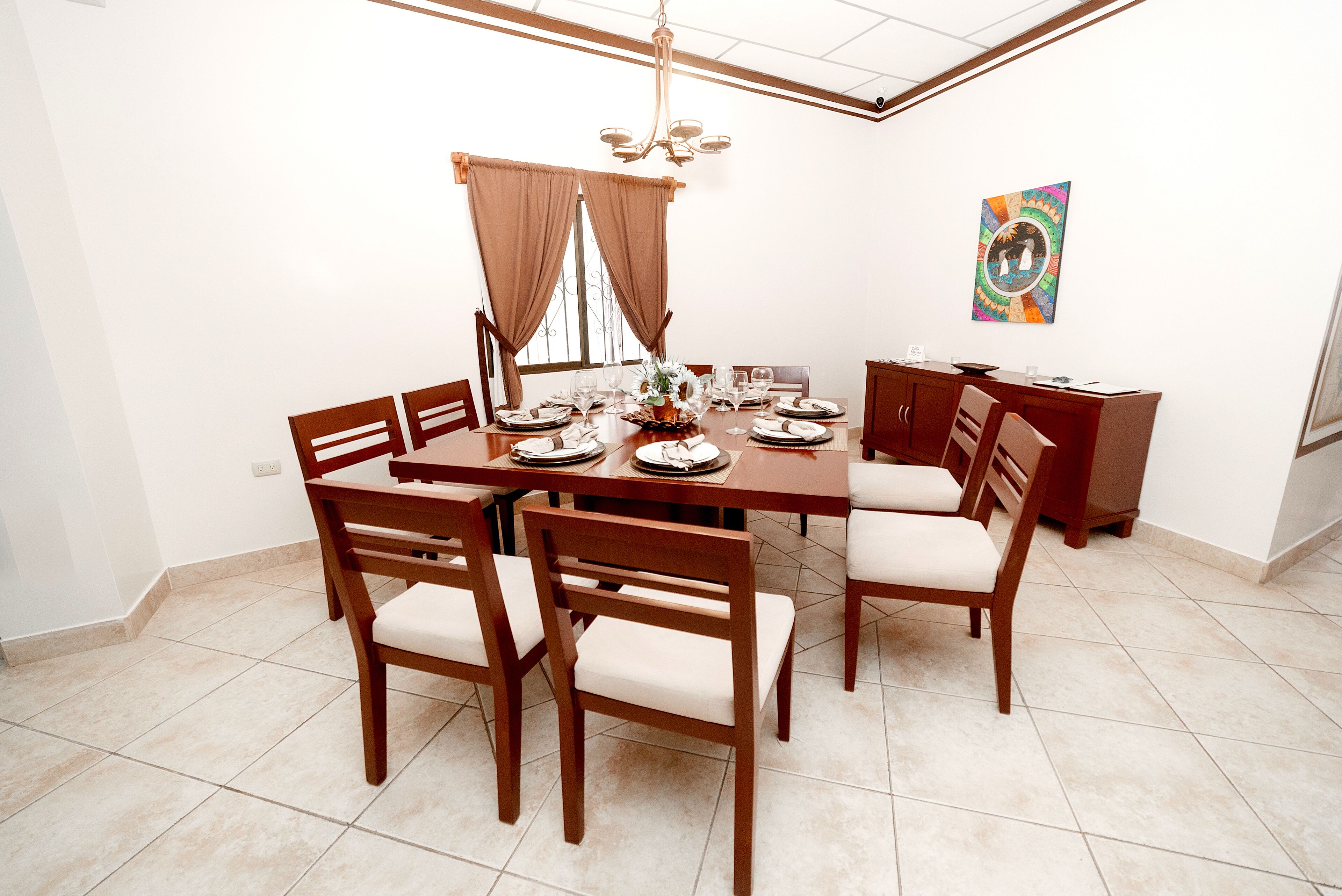 A dining area with a wooden table set for six, surrounded by chairs, a chandelier, and a decorative wall piece.