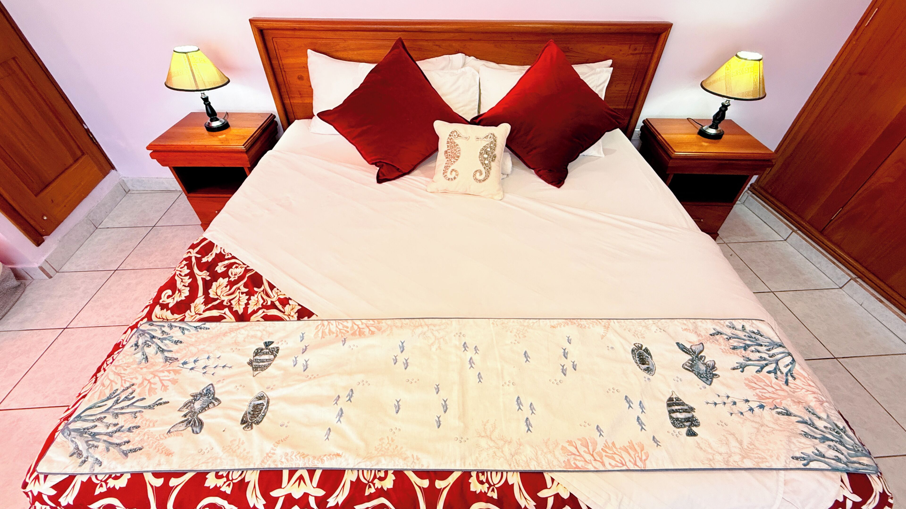 A neatly made bed with a red and white patterned bedspread, two red pillows, and a decorative pillow with a seahorse design. There are two bedside tables with lamps, and the room has a wooden headboard.