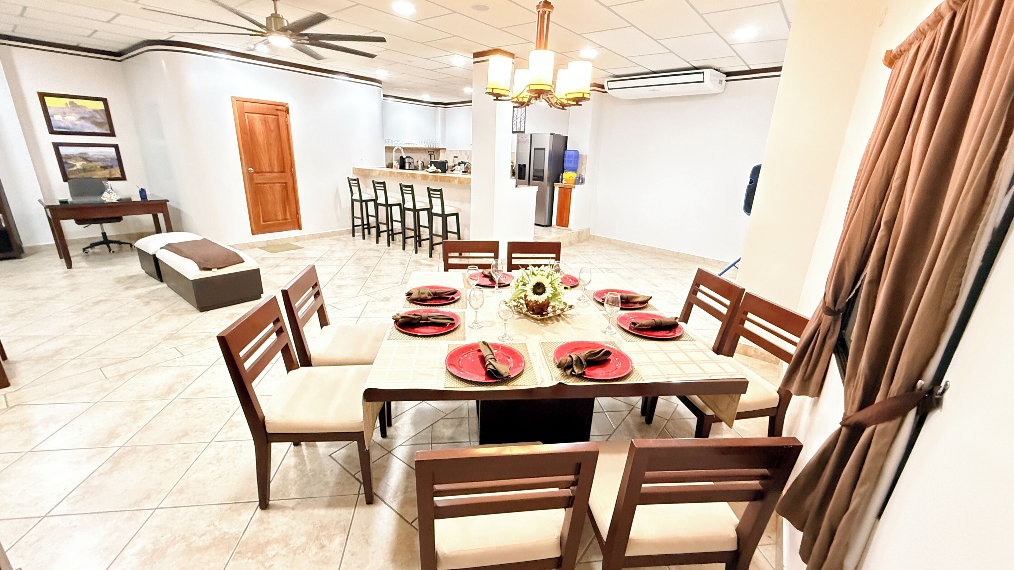 A dining area with a round table set for six, surrounded by chairs, with a kitchen in the background.