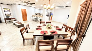 A dining area with a round table set for six, surrounded by chairs, with a kitchen in the background.
