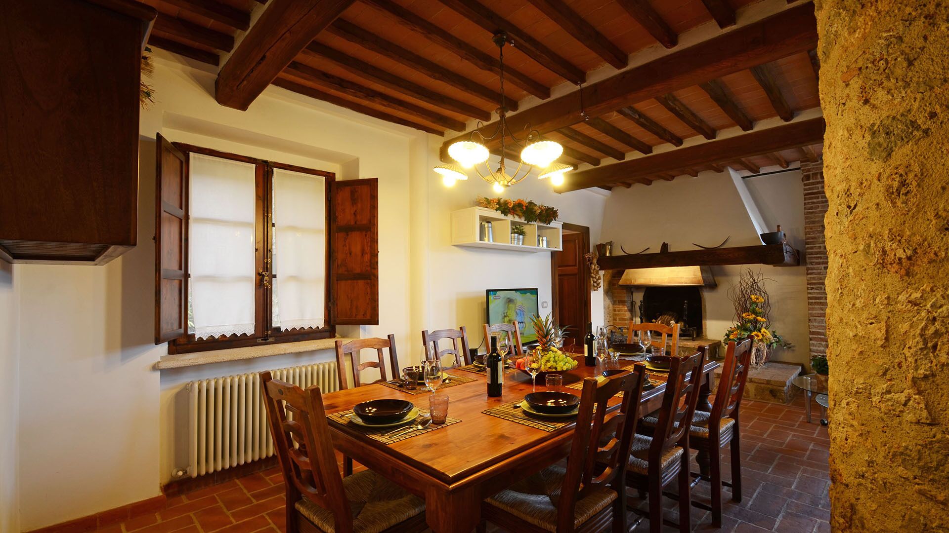 A dining area with a wooden table set for four, flanked by wooden chairs. A chandelier hangs from the ceiling, and a brick fireplace is visible in the background.