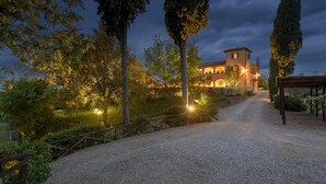 Un sentiero sterrato conduce a un edificio con una torre, immerso tra alberi e illuminato da luci calde.
