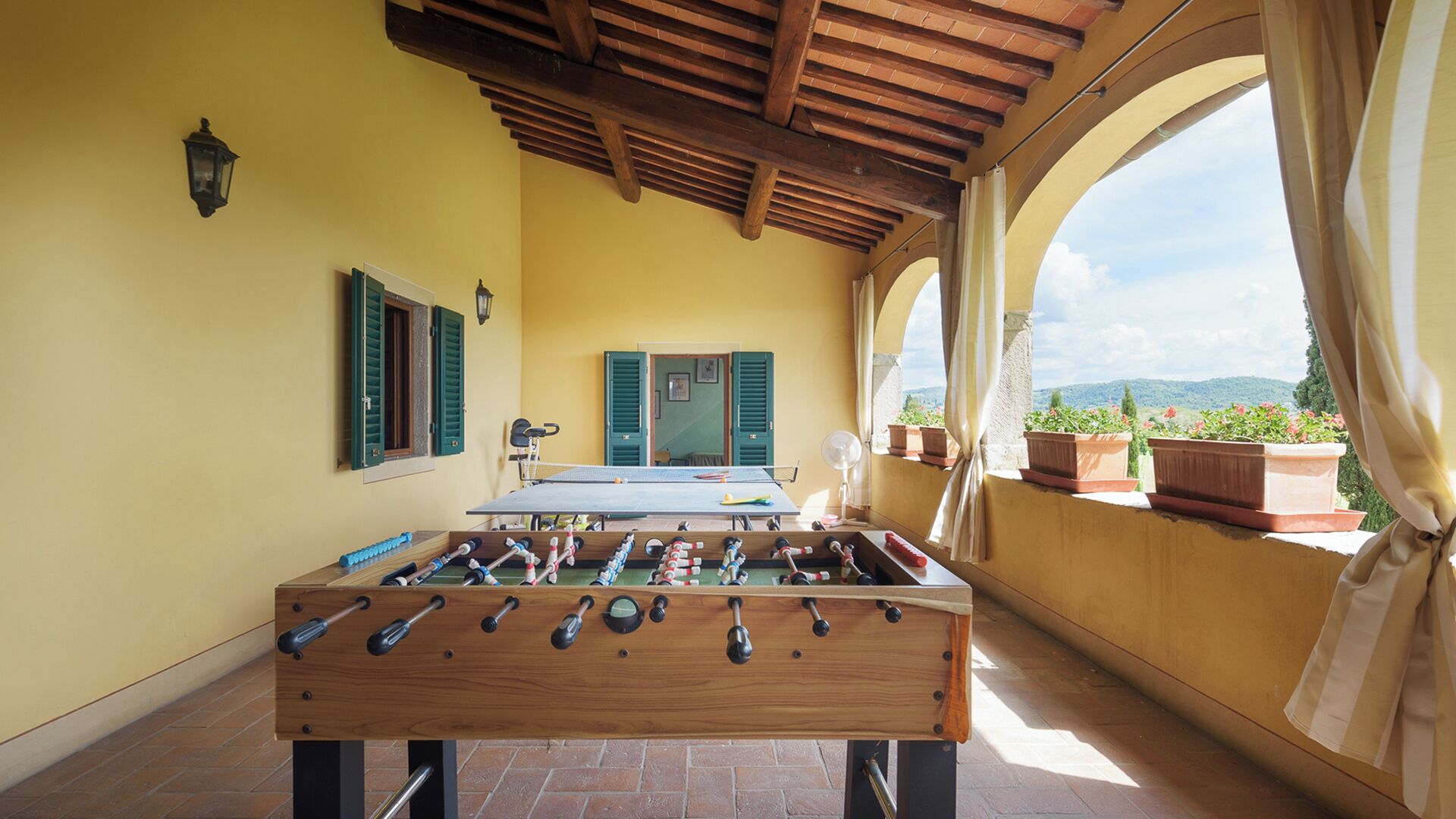 A foosball table in a room with yellow walls, wooden ceiling beams, and a view through an arched window.