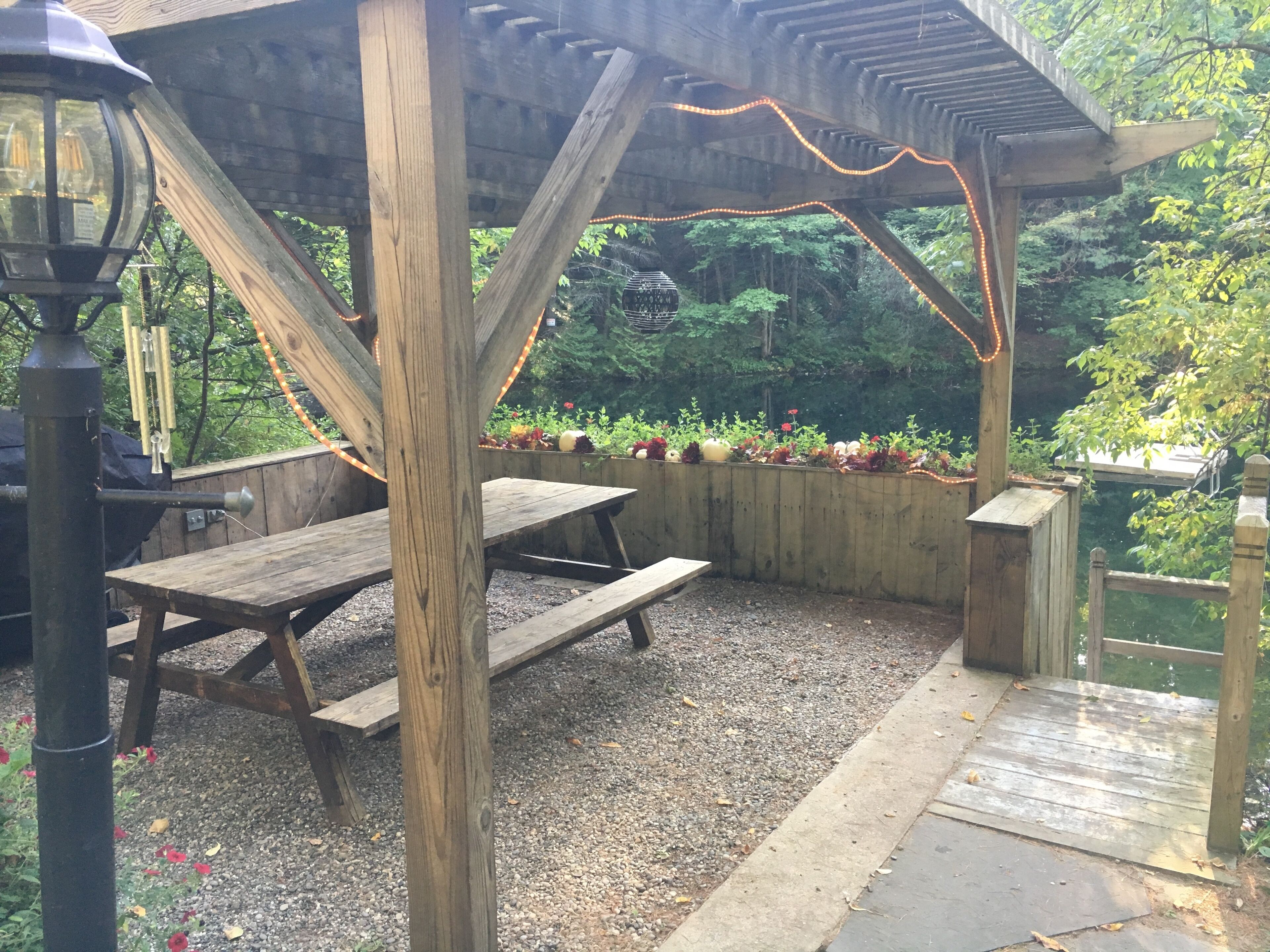 Eine Holzpergola mit Lichterketten, Picknicktischen und einem Garten mit verschiedenen Pflanzen und Blumen.