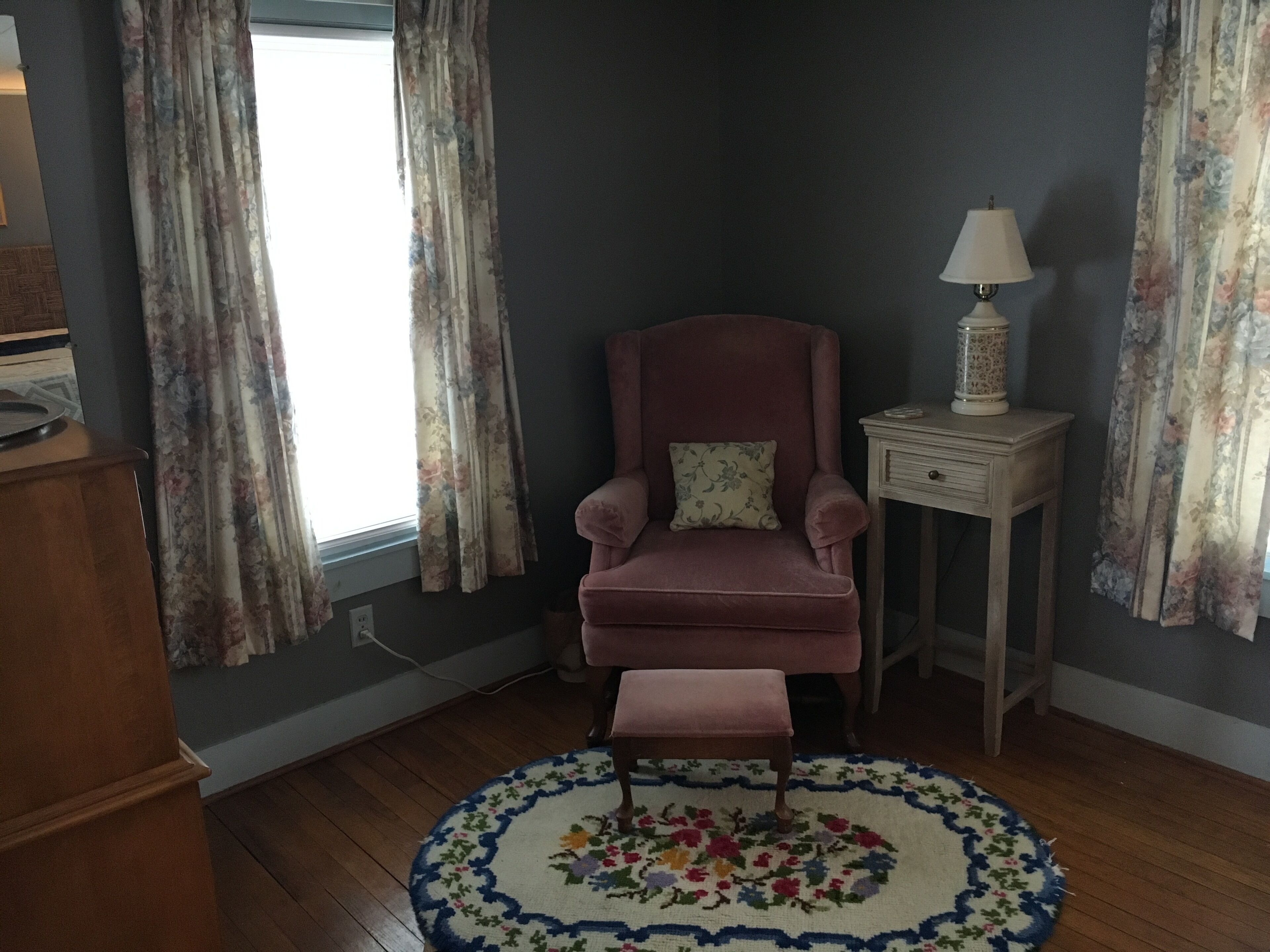 Une pièce avec un mur de couleur sombre, un fauteuil rose orné d’un coussin à motifs floraux, une petite table d’appoint en bois avec une lampe, et un tapis rond à motifs floraux.