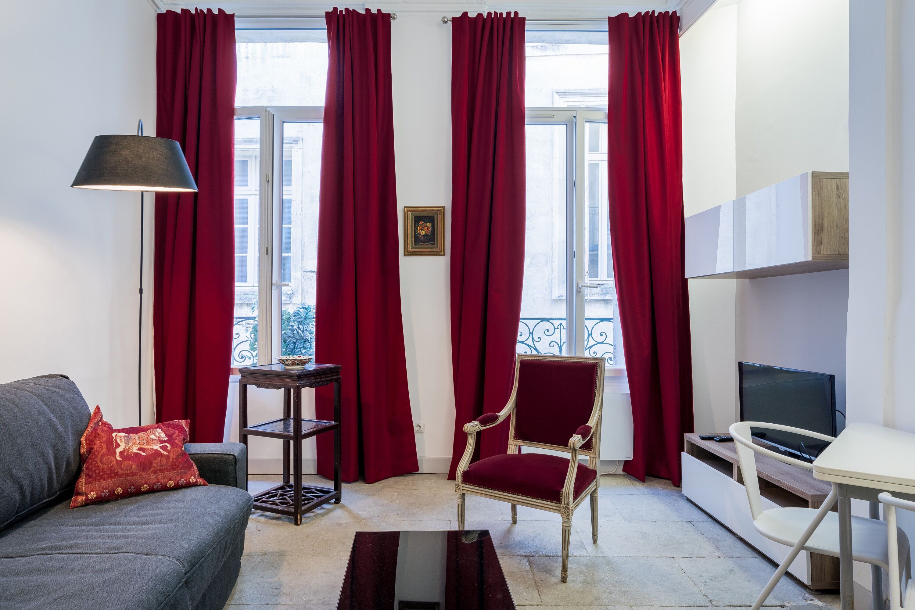 A living room with a sofa, a red chair, a small table, a TV, and a window with red curtains.