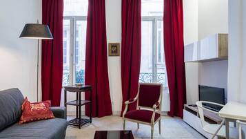 A living room with a sofa, a red chair, a small table, a TV, and a window with red curtains.