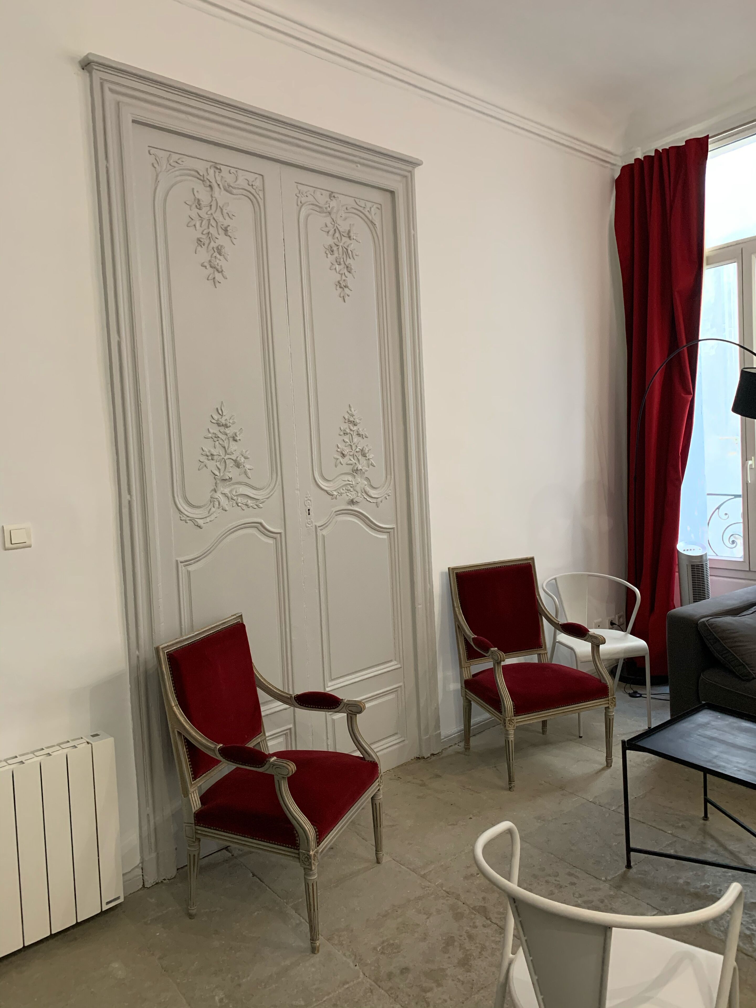 A room with a large white wardrobe, two red upholstered chairs, a white chair, a grey sofa, and a small table.