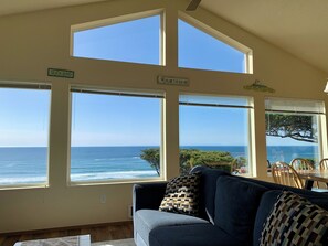A room with a large window overlooking the ocean, a couch with patterned cushions, and a dining table with chairs.