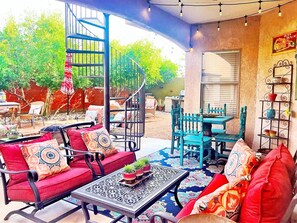 Ein Patio mit einer Wendeltreppe, Sitzgelegenheiten im Freien mit roten und blauen Stühlen, einem Tisch mit gemustertem Tischläufer und einem dekorativen Regal mit Blumentöpfen.