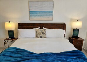 Ein Bett mit weißen Leinentüchern, einer blauen Decke, zwei Nachttischen mit Lampen und einem Bild einer Strandansicht über dem Bett.