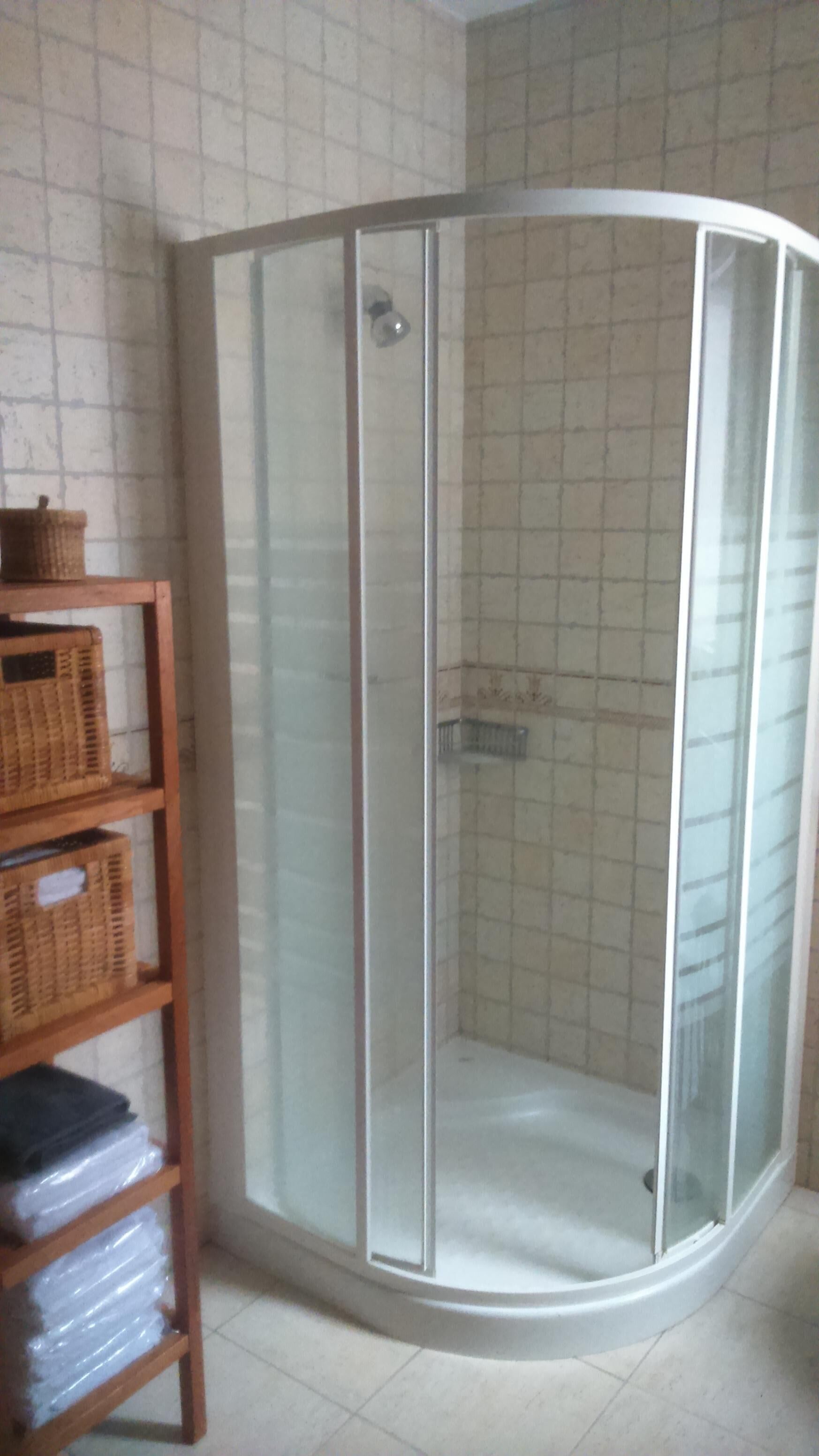 A corner shower with a glass door, a wooden shelf with baskets, and tiled walls.
