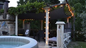 Ein Außenbereich mit Whirlpool, einer Steinwand, einem mit Lichterketten geschmückten Pergola und einer Holzterrasse mit Gartenmöbeln.