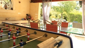 Une table de baby-foot dans une pièce où se trouvent une table et des chaises en verre, une fenêtre avec des rideaux et une vue sur la verdure à l’extérieur.