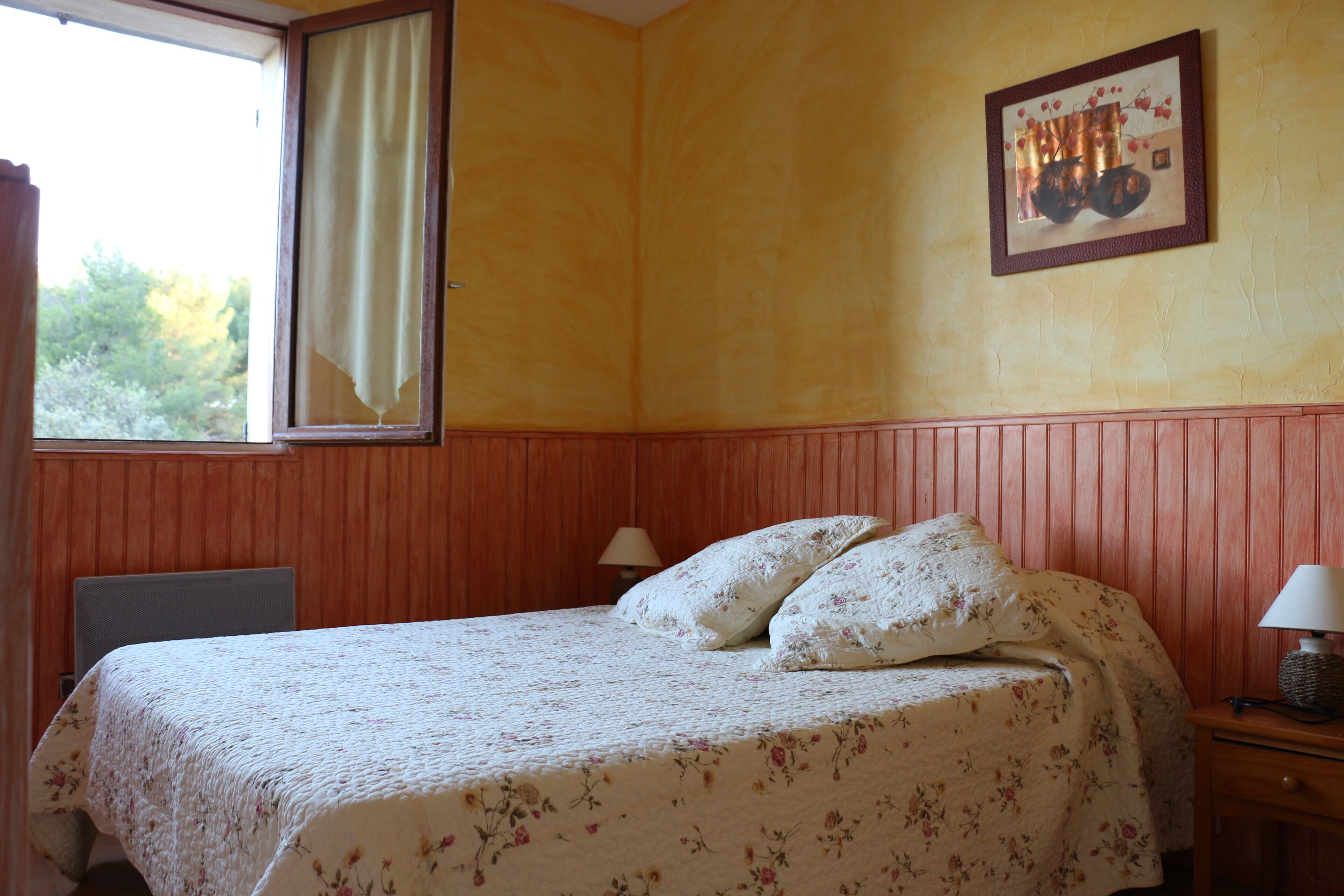 Une chambre à coucher comprenant un lit, une table de chevet, une lampe, une fenêtre avec des rideaux et un tableau accroché au mur.