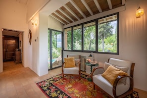 A hallway with a wooden ceiling, a large window with a view of greenery, two white chairs with wooden frames, and a patterned rug.